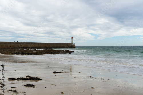 Vue sur le môle et le phare du Raoulic, avec en premier plan du sable mouillé, des rochers et un océan de couleur émeraude, sous un ciel couvert en Bretagne.