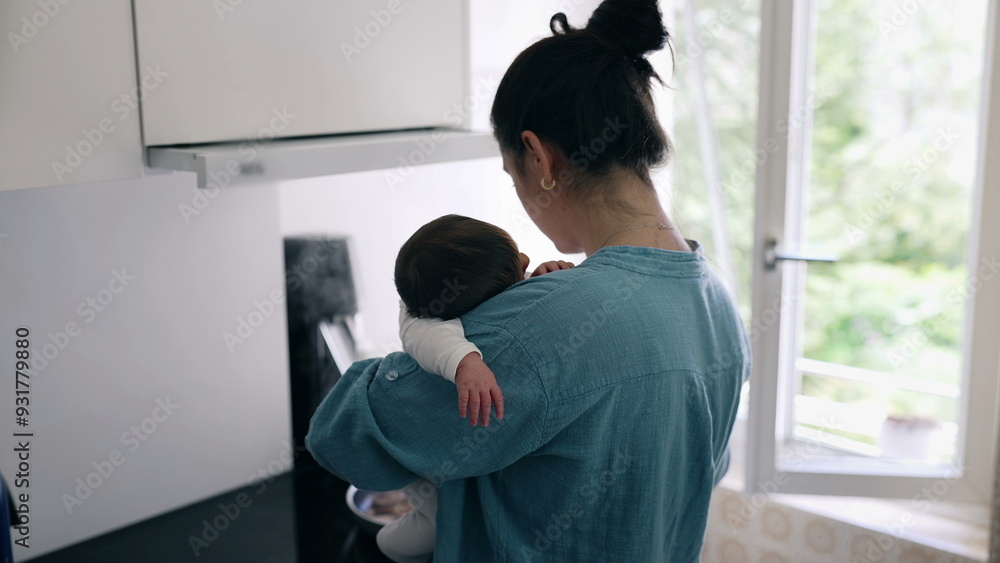 Mother in a blue shirt holding a baby while cooking in a modern kitchen with natural light coming through the window, showcasing multitasking and nurturing care in a cozy home environment Mother in a blue shirt holding a baby while cooking in a modern kitchen with natural light coming through the window, showcasing multitasking and nurturing care in a cozy home environment
