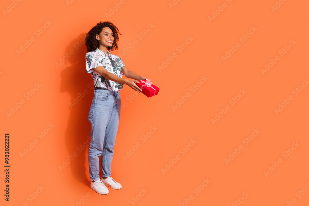 Photo of young beautiful happy girl curly hairstyle holding birthday box looking and giving surprise isolated on orange color background Photo of young beautiful happy girl curly hairstyle holding birthday box looking and giving surprise isolated on orange color background