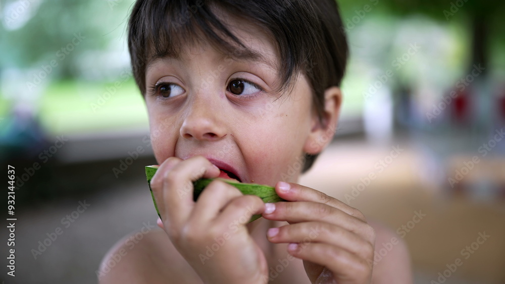 Fototapeta premium Boy savoring watermelon slice on a warm day, park setting, casual summer moment, natural backdrop, childhood joy, fresh fruit snack, healthy eating, happy and relaxed, playful expression