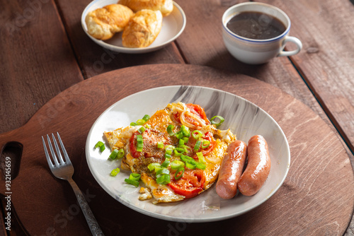 Eggs with tomatoes and fried sausage in a plate on a wooden table of coffee and croissants on a round stand