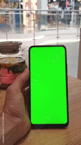 In a food court in the mall at a table with tea, the hand holds vertically a smartphone with a green screen. Vertical Video Template for Advertising and Apps.