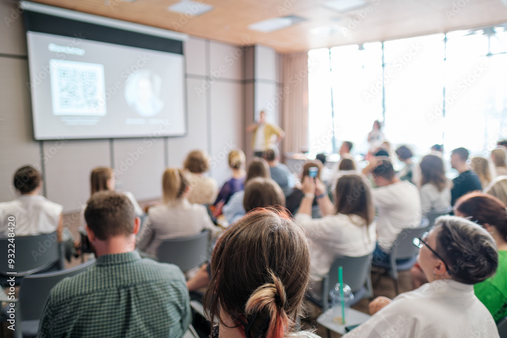 Conference attendees focusing on speaker during presentation with projected slides. Engaging business meeting atmosphere with active participation. Conference attendees focusing on speaker during presentation with projected slides. Engaging business meeting atmosphere with active participation.