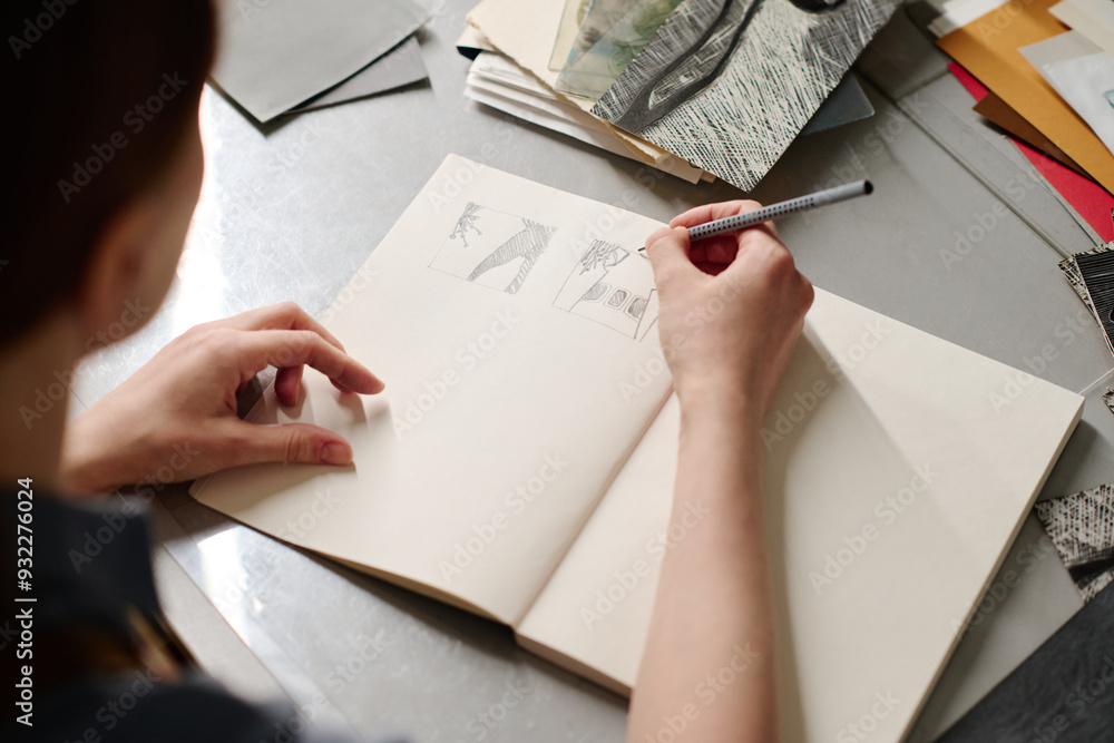 Fototapeta premium Young craftswoman with pencil sitting by workplace with artwork samples and making new sketches on page of open notepad