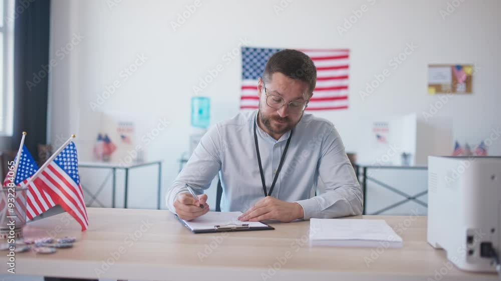 Handsome Caucasian male with beard sitting at desk in large office. Writing something with pen on piece of paper while checking pile of blanks next to him. American flags everywhere in room. Elections Handsome Caucasian male with beard sitting at desk in large office. Writing something with pen on piece of paper while checking pile of blanks next to him. American flags everywhere in room. Elections
