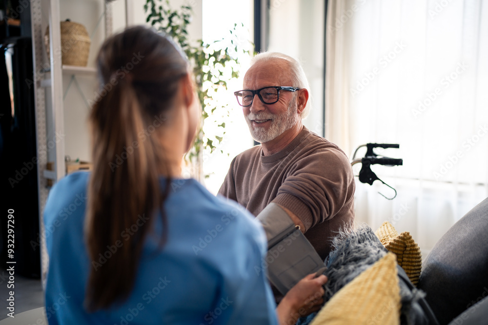 Obraz premium A smiling female nurse in blue scrubs checks the blood pressure of an elderly man with white hair and glasses in his home, reflecting professional home healthcare services.