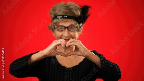 Funny portrait of elderly old woman toothless, giving heart gesture with hands isolated on red background wearing wig, glasses