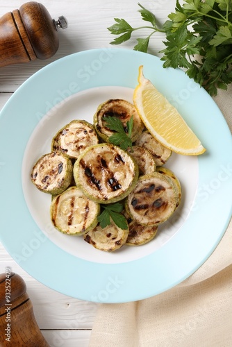 Tasty grilled zucchini slices with lemon and parsley on white wooden table, flat lay