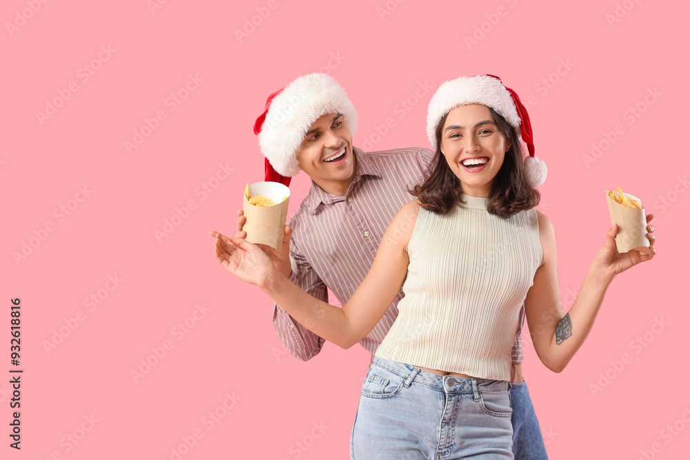 Young couple in Santa hats with french fries on pink background Young couple in Santa hats with french fries on pink background