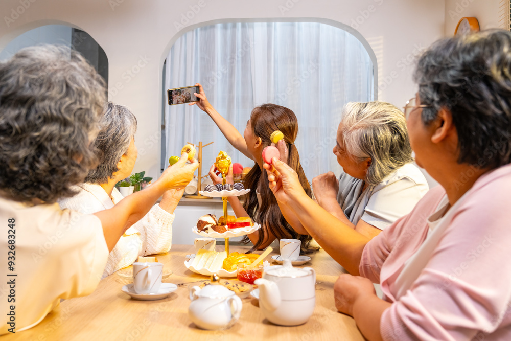 Group of Happy Asian senior women having meeting party afternoon tea at home. Daughter using mobile phone taking selfie with elderly retired mother and friends during drinking tea and eating bakery Group of Happy Asian senior women having meeting party afternoon tea at home. Daughter using mobile phone taking selfie with elderly retired mother and friends during drinking tea and eating bakery