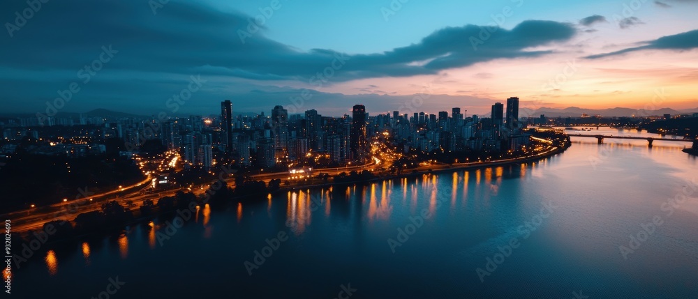Naklejka premium Modern city skyline at dusk with warm lights reflecting on a calm river. The scene captures the transition from day to night, with soft clouds adding to the atmosphere.