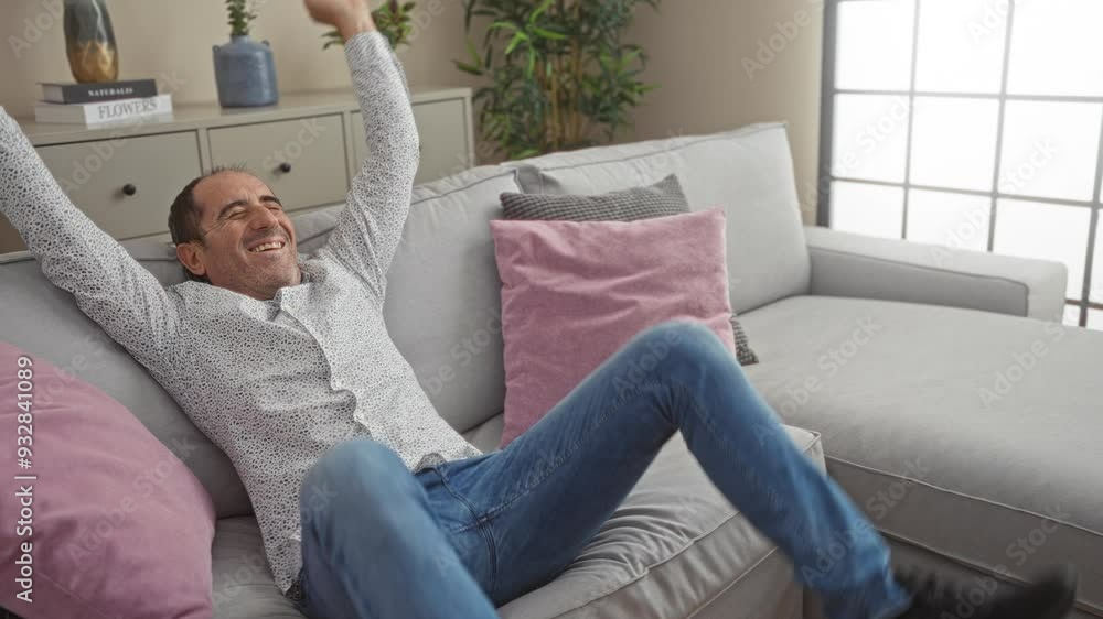 Middle-aged man celebrating indoors at home sitting on a sofa in the living room with arms raised and fingers crossed wearing jeans and a patterned shirt in a cheerful mood Middle-aged man celebrating indoors at home sitting on a sofa in the living room with arms raised and fingers crossed wearing jeans and a patterned shirt in a cheerful mood
