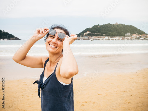 Portrait of a cheerful woman on the beach, adjusting her sunglasses and smiling, with a scenic view of the coastline and hills in the background.