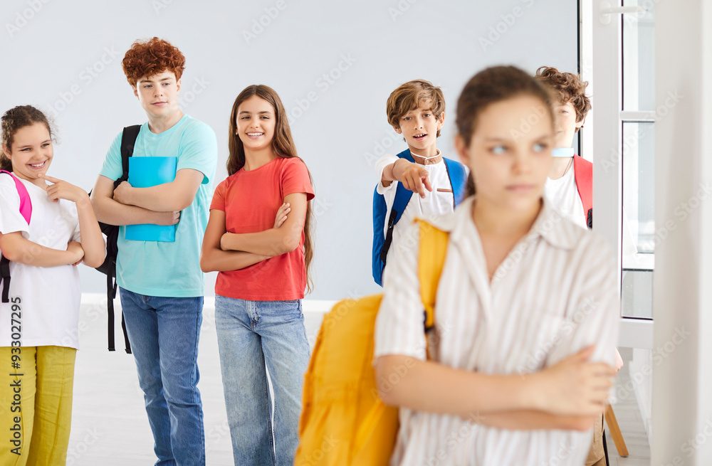 Outcast sad female teenager suffering from bullying, standing alone with group of classmates laughing behind her back in school corridor. Stressed victim girl feeling lonely and excluded at school. Outcast sad female teenager suffering from bullying, standing alone with group of classmates laughing behind her back in school corridor. Stressed victim girl feeling lonely and excluded at school.