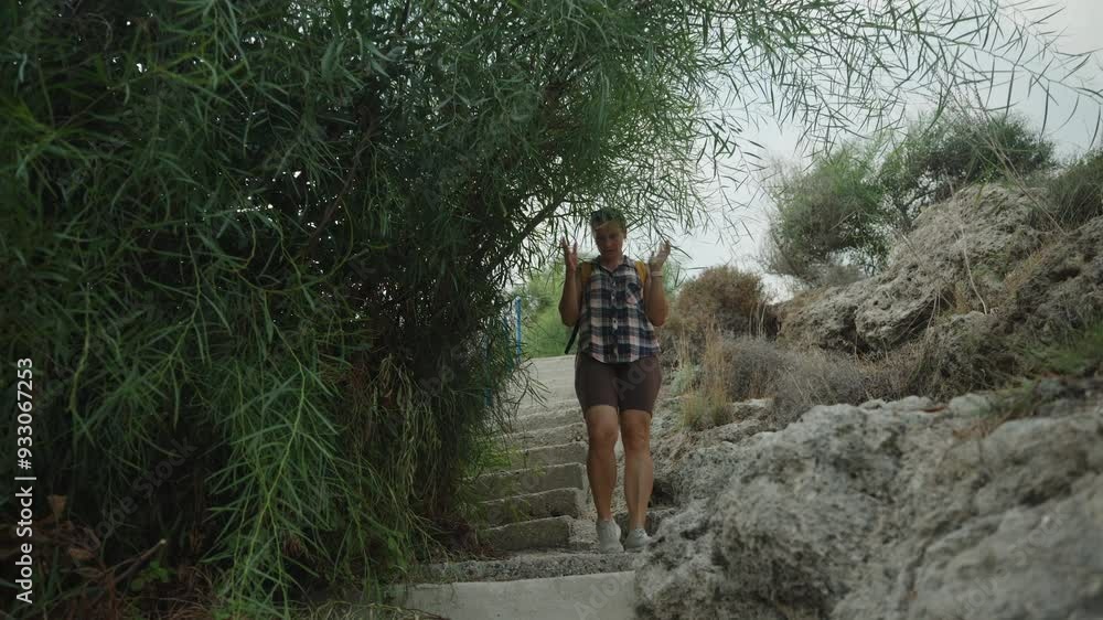 A Woman Carefully Descends Stone Steps Surrounded by Lush Greenery. She Gently Pushes Branches Aside While Navigating the Narrow Path. A Woman Carefully Descends Stone Steps Surrounded by Lush Greenery. She Gently Pushes Branches Aside While Navigating the Narrow Path.
