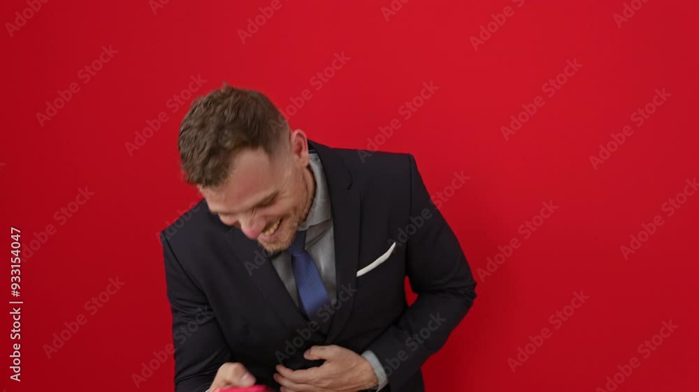 Young hispanic man in suit, laughing out loud while having fun with a funny joke on his smartphone, smiling against an isolated red background
