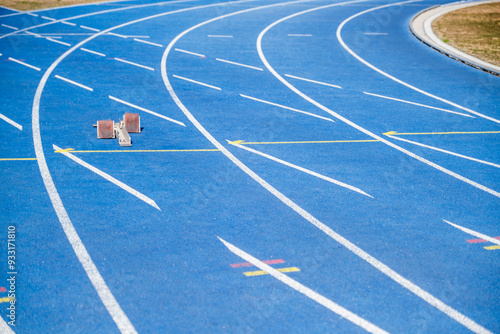 Vibrant blue running track with starting blocks, empty stadium, and textured background. Athletes prepare to race on a sunny day