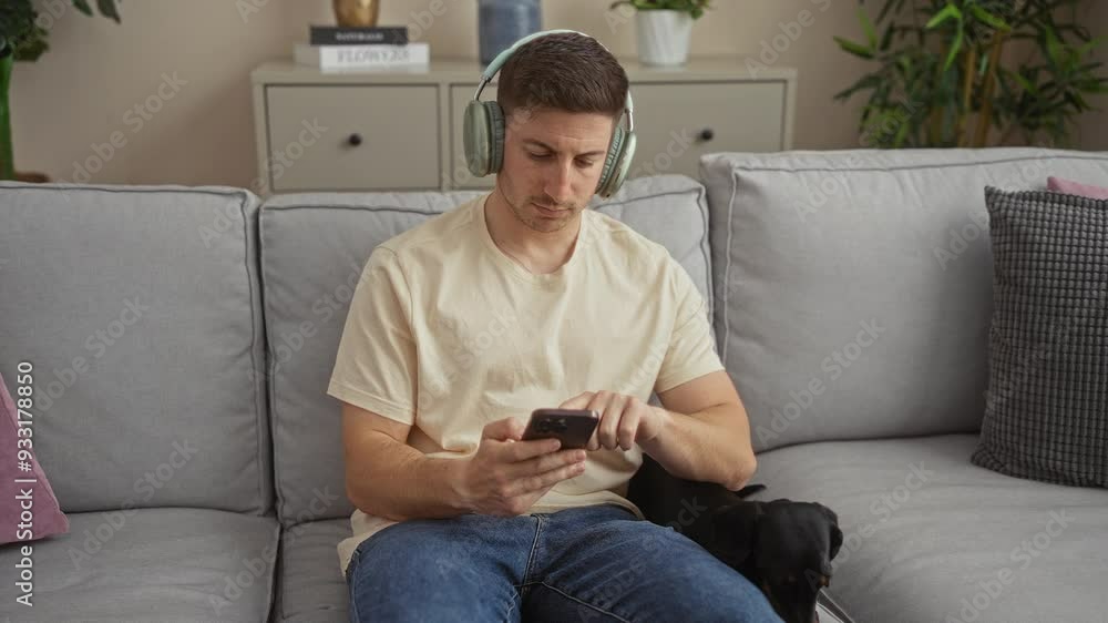 Young hispanic man wearing headphones sitting in a living room with a dachshund, using his smartphone. Young hispanic man wearing headphones sitting in a living room with a dachshund, using his smartphone.