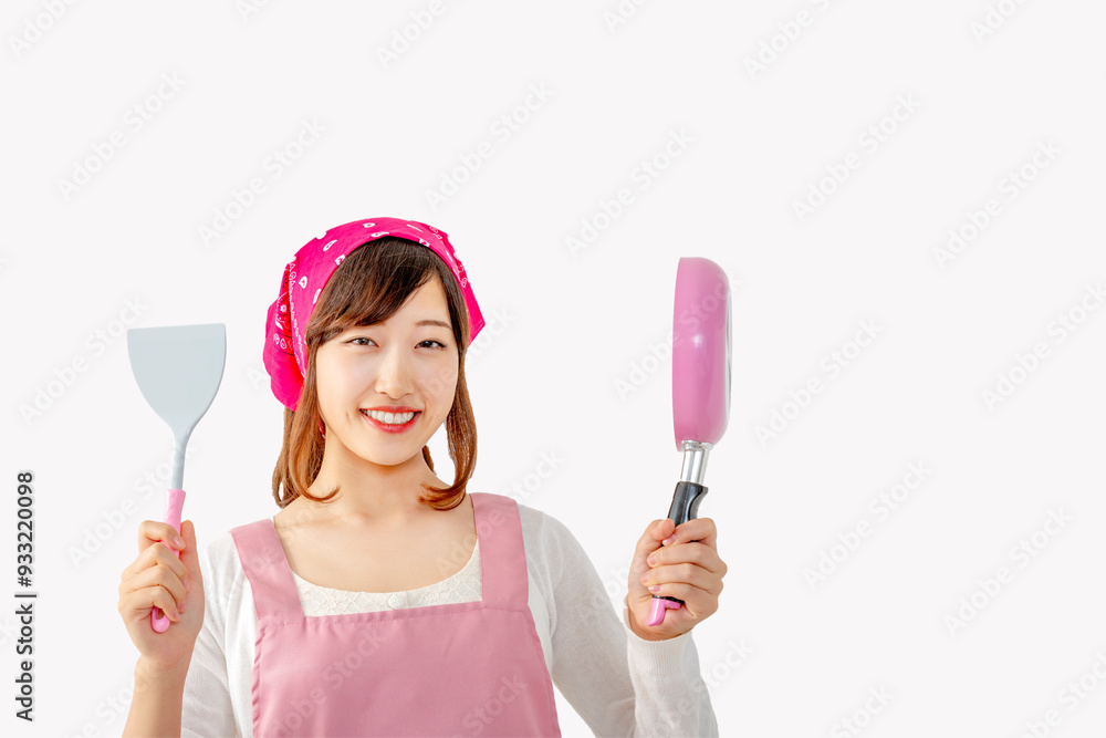 白背景の前でフライ返しとフライパンを持ちヤル気をアピールする、笑顔の若い女性1人