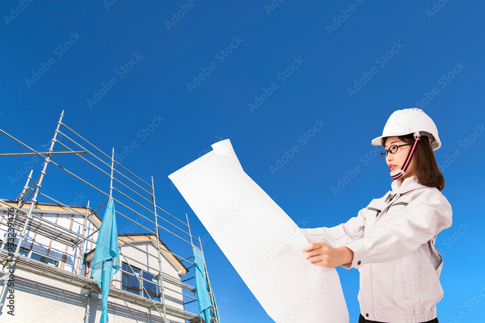 青空と戸建て工事現場を背景に立ち、図面を見る作業着を着た若い女性