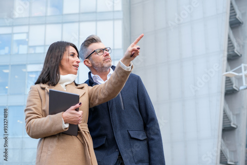 Confident business duo discussing strategy outside office building in city, using tablet for collaboration. Modern architecture reflects forward-thinking nature