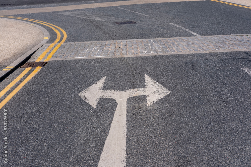 A close-up of a road surface with bold white arrows marking directions for left and right turns, accompanied by yellow lines and a paved crosswalk in a city setting. A close-up of a road surface with bold white arrows marking directions for left and right turns, accompanied by yellow lines and a paved crosswalk in a city setting.