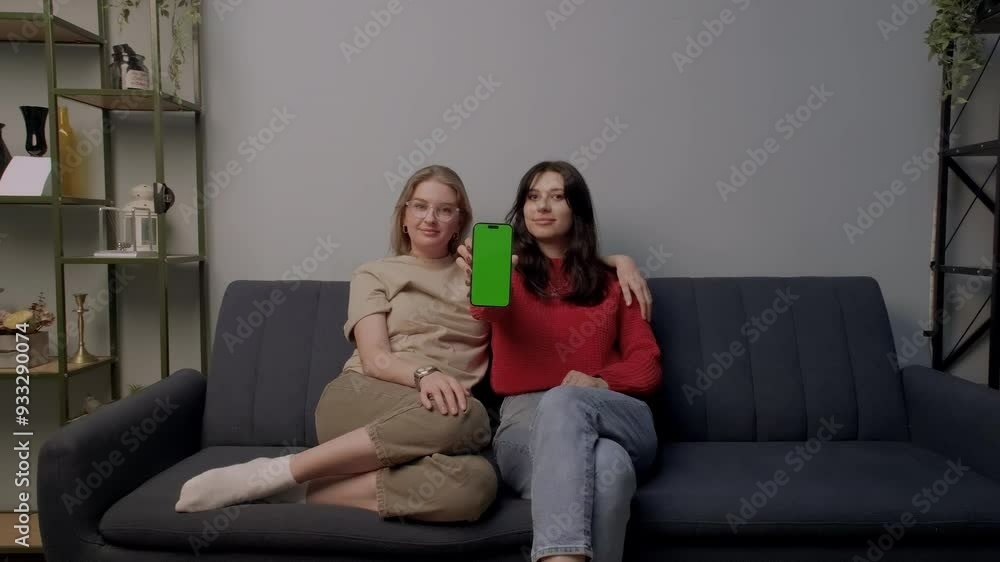 Young lesbian LGBTQ couple sitting on sofa and showing phone with green screen Young lesbian LGBTQ couple sitting on sofa and showing phone with green screen