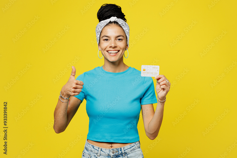 Young fun Latin woman she wears blue top t-shirt headband casual clothes hold in hand mock up of credit bank card show thumb up isolated on plain yellow background studio portrait. Lifestyle concept. Young fun Latin woman she wears blue top t-shirt headband casual clothes hold in hand mock up of credit bank card show thumb up isolated on plain yellow background studio portrait. Lifestyle concept.