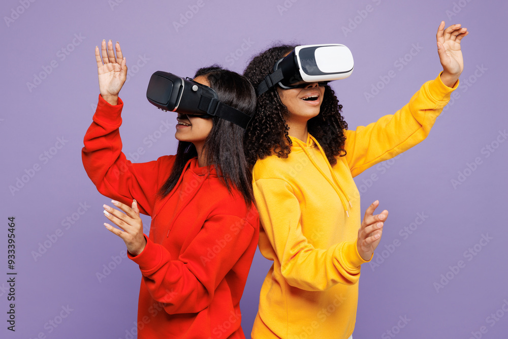 Obraz premium Sideways two young happy little kid teen girls women they wearing red yellow hoodie casual clothes watching in vr headset pc gadget isolated on plain pastel light purple background. Lifestyle concept.
