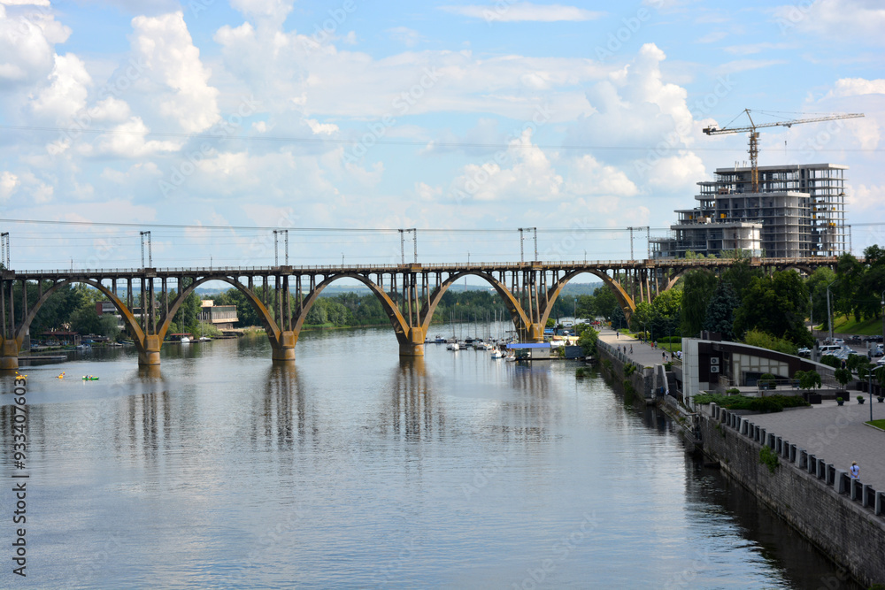 Obraz premium Beautiful view of the wide and long blue Dnipro River. Landscapes, a gray brick embankment with green trees, new construction sites and a beautiful bridge in the city of Dnipro.