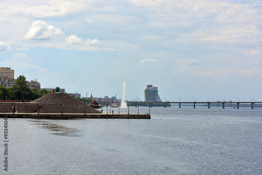 Beautiful view of the wide and long blue Dnipro River. Landscapes, a gray brick embankment with green trees, new construction sites and a beautiful bridge in the city of Dnipro. Beautiful view of the wide and long blue Dnipro River. Landscapes, a gray brick embankment with green trees, new construction sites and a beautiful bridge in the city of Dnipro.