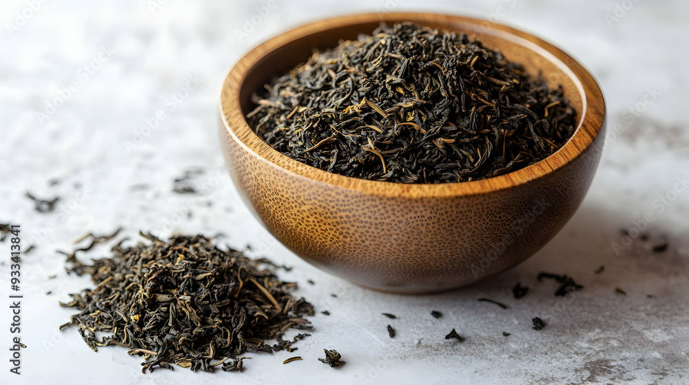 There is a wooden bowl filled with loose tea leaves next to a small pile of the same tea leaves on a white table. There is a wooden bowl filled with loose tea leaves next to a small pile of the same tea leaves on a white table.