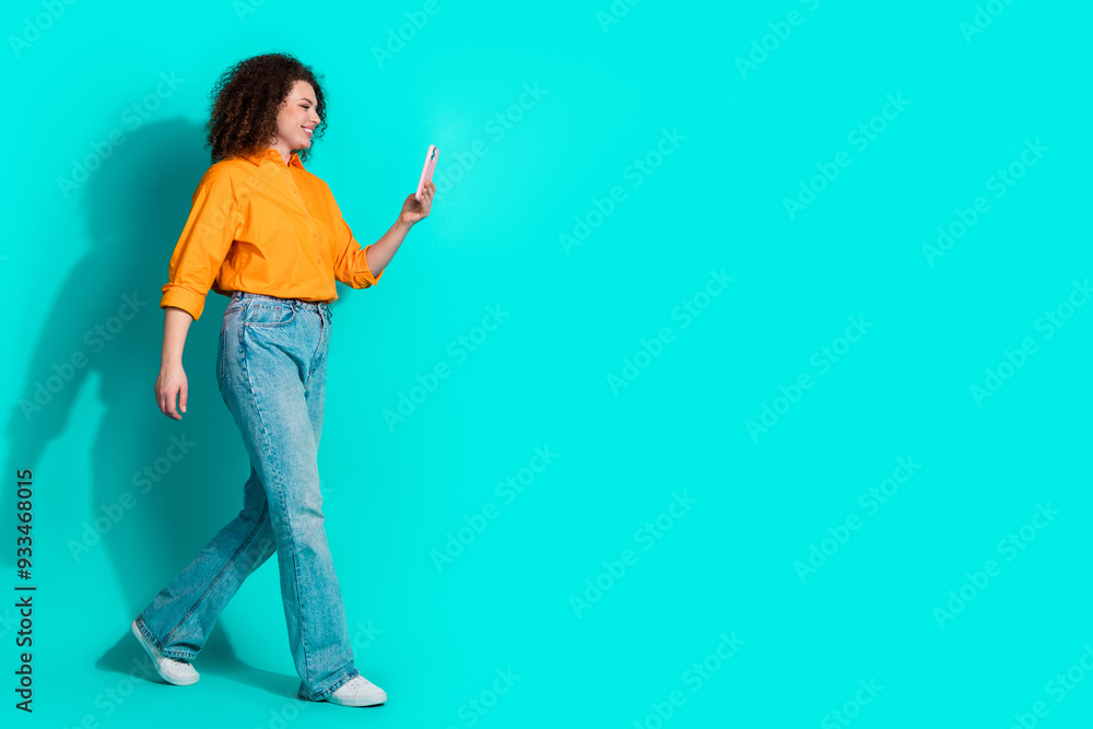 Full size photo of positive optimistic woman dressed orange shirt look at smartphone go empty space isolated on teal color background Full size photo of positive optimistic woman dressed orange shirt look at smartphone go empty space isolated on teal color background