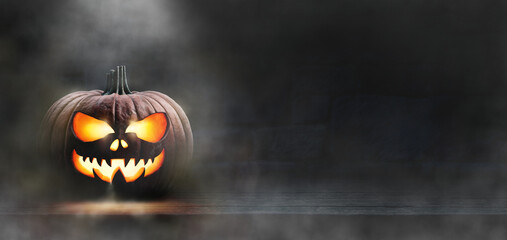 A scary spooky glowing carved halloween pumpkin lantern, Jack O' Lanterns, on the left of a wooden product display bench on a scary halloween night with a dark stone wall background lit from above.