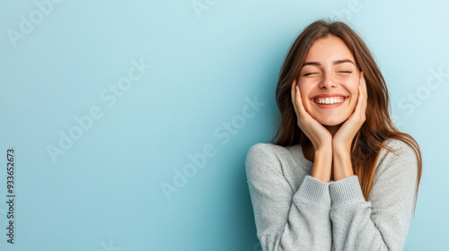 Young woman smiling joyfully with her hands on her cheeks, expressing delight and happiness against a soft blue background. photo