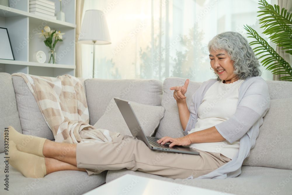 Elderly woman with curly gray hair is comfortably reclining on a light-colored sofa in a bright, modern living room she is engaged in a pleasant video call or conversation. Elderly woman with curly gray hair is comfortably reclining on a light-colored sofa in a bright, modern living room she is engaged in a pleasant video call or conversation.