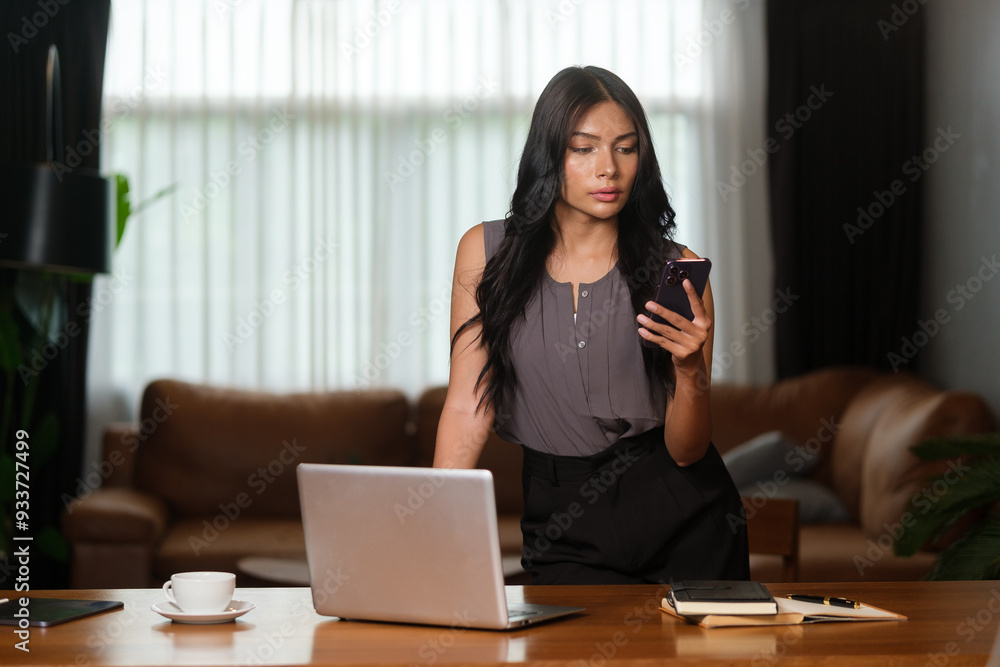 Young professional woman is standing at a wooden desk in a modern office setting, focused on her smartphone. Young professional woman is standing at a wooden desk in a modern office setting, focused on her smartphone.