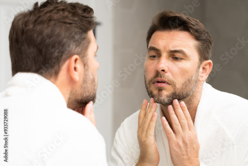 a man in a bathrobe in the bathroom looking at his wrinkles and pimples on his face. A man is looking at his reflection in a mirror. A man is looking at his reflection in a mirror. 