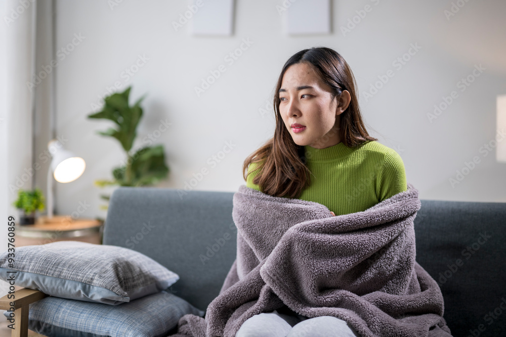 Young woman feeling cold sitting on sofa covering herself with blanket at home Young woman feeling cold sitting on sofa covering herself with blanket at home