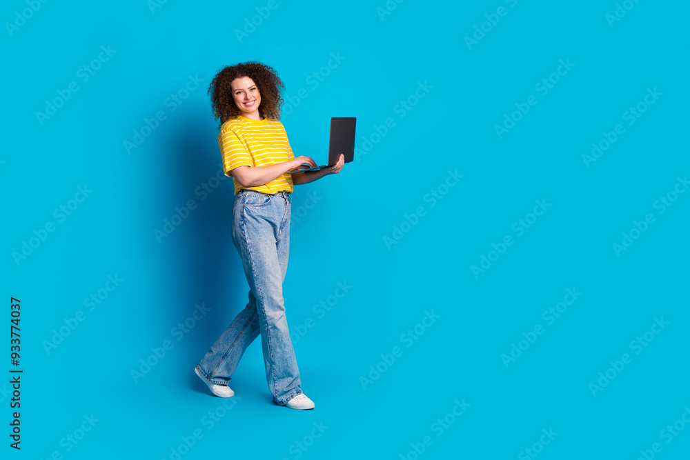 Full size photo of pretty young girl walking hold netbook wear trendy striped yellow outfit isolated on blue color background Full size photo of pretty young girl walking hold netbook wear trendy striped yellow outfit isolated on blue color background