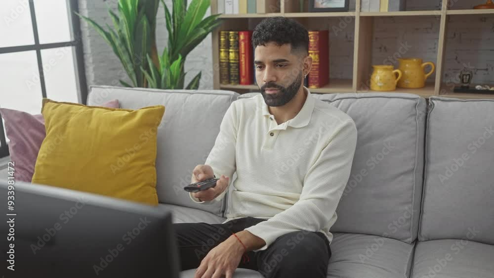 Handsome young hispanic man with beard sitting on grey couch in living room holding remote Handsome young hispanic man with beard sitting on grey couch in living room holding remote