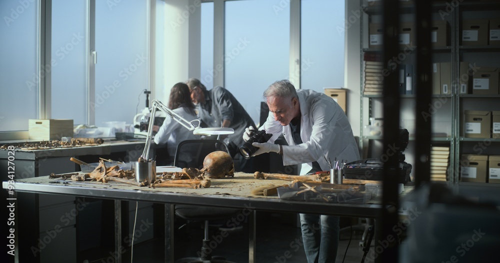Modern archaeological laboratory: Senior archaeologist takes picture of fossil remains of prehistoric man skull with professional camera. Female scientist and archaeologist work in the background. Modern archaeological laboratory: Senior archaeologist takes picture of fossil remains of prehistoric man skull with professional camera. Female scientist and archaeologist work in the background.