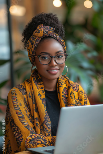 Joyful dark-skinned female blogger typing at a cozy cafe urrounded by plants during a sunny afternoon photo