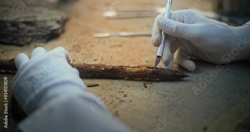 Senior archaeologist in gloves cleans fossil ancient wooden spear using professional tools. Scientist studies weapon remains of extinct human civilization, works in archaeological laboratory. Close Up