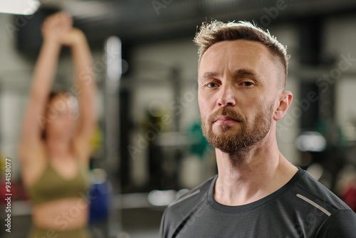 Portrait of confident male coach with trimmed beard looking at camera, female coach warming up in background