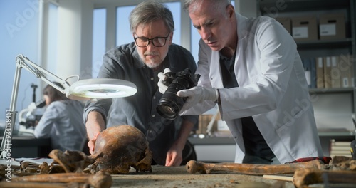 Canvas Print Senior paleoanthropologist takes photo of fossil remains of prehistoric human skull with colleague archaeologist in archaeological laboratory using camera
