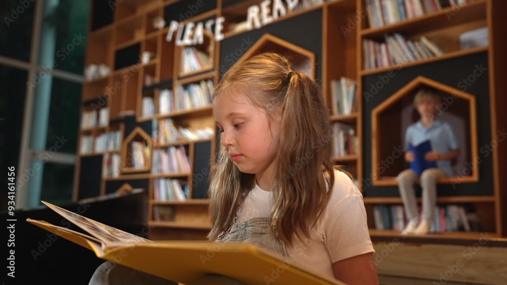 Young smart caucasian girl picking reading a book while sitting at library. Clever child learning, studying, open a books at library. Attractive kid turning page with blurring background. Erudition. Young smart caucasian girl picking reading a book while sitting at library. Clever child learning, studying, open a books at library. Attractive kid turning page with blurring background. Erudition.