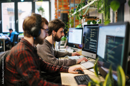 Focused young programmers coding at computers in a modern office with green plants background. Concepts of technology, teamwork, and productivity in a creative work environment.