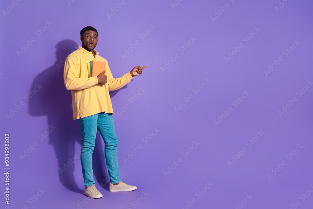 Full length photo of handsome young guy hold books point empty space dressed stylish yellow garment isolated on violet color background Full length photo of handsome young guy hold books point empty space dressed stylish yellow garment isolated on violet color background