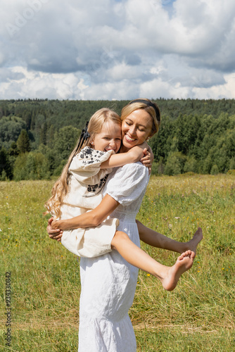 young mother holds her teenage daughter in her arms and hugs her smiling they stand in the middle of the grass in the meadow, tenderness and childhood